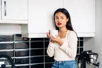 Жадібна етнічна жінка в повсякденному одязі сидить на стільниці з чашкою кави, відпочиваючи вдома — стокове фото