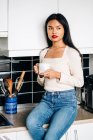 Wistful ethnic female in casual clothes sitting on countertop with cup of coffee while resting at home — Stock Photo