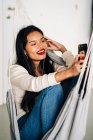 Glückliche ethnische Frau mit zahmem Lächeln sitzt mit Kopfhörern in der Hängematte und hat Videoanrufe auf dem Handy — Stockfoto