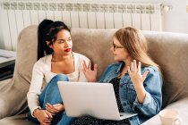 Happy woman with laptop sitting on sofa and gesticulating while communicating with pensive ethnic female friend — Stock Photo