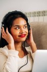 Side view of focused ethnic female lying on sofa with headphones working on modern laptop — Stock Photo