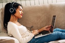 Вид збоку зосередженої етнічної жінки, що лежить на дивані з навушниками, що працюють на сучасному ноутбуці — стокове фото