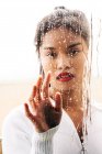 Through glass of pensive ethnic female with vivid lips looking at camera while touching drops — Stock Photo
