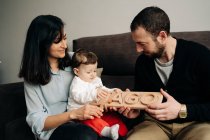 Feliz joven multirracial padres sentados en el sofá y jugando con adorable pequeño hijo sosteniendo juguete de madera en casa - foto de stock