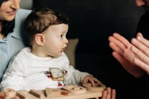 Crop unrecognizable young father clasping hands while playing with adorable little son sitting on knees of ethnic mother with wooden toy in hands — Photo de stock