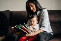 Обрізати молоду етнічну матір в повсякденному одязі, сидячи на дивані і читаючи книгу для чарівного маленького сина — стокове фото