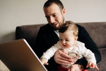 Young bearded man hugging little son while sitting on comfortable sofa and watching cartoon on laptop at home — Fotografia de Stock