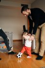 Funny little boy kicking ball while playing with young father and crop unrecognizable mother at home — Photo de stock