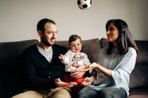 Amorosos padres jóvenes sentados en el sofá y abrazando a su pequeño hijo lindo jugando con la pelota en casa - foto de stock