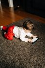 Сверху содержание маленького мальчика лежит на пушистом ковре и смотрит смешное видео на мобильном телефоне в светлой гостиной — стоковое фото