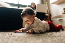Contenido pequeño bebé niño acostado en una alfombra esponjosa y viendo videos divertidos en el teléfono móvil en la sala de estar de luz - foto de stock
