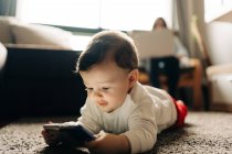 Content little baby boy lying on fluffy carpet and watching funny video on mobile phone in light living room — Stock Photo