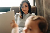 Zufriedene junge Mutter sitzt auf bequemem Stuhl und blättert im Netbook neben entzückendem kleinen Sohn — Stockfoto