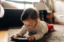 Content little baby boy lying on fluffy carpet and watching funny video on mobile phone in light living room — Fotografia de Stock