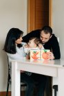 Позитивные молодые родители и очаровательный маленький сын, сидящий за столом и читающий детскую книгу вместе — стоковое фото