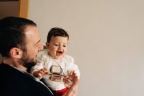 Alegre joven padre celebración adorable divertido hijo en manos mientras se divierten juntos en sala de luz - foto de stock