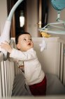 From above adorable little boy standing in comfy crib and playing with white gumshoe — Photo de stock