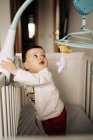 From above adorable little boy standing in comfy crib and playing with white gumshoe — Fotografia de Stock