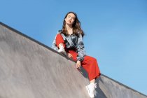 Низький кут жіночого підлітка в модному одязі і камерах дивиться на камеру з бетонного паркану в спині, освітленій блакитним прозорим небом — стокове фото