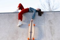 Low angle side view of tired female in trendy outfit with longboard lying in concrete skate park on sunny day — Photo de stock