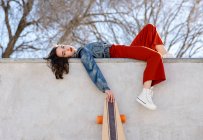 Low angle side view of tired female in trendy outfit with longboard lying in concrete skate park on sunny day looking at camera — Photo de stock