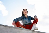 Niedriger Winkel einer Teenagerin in trendigen Klamotten und Gummischuhen, die von einem Betonzaun wegschaut, im Gegenlicht mit blauem Himmel — Stockfoto