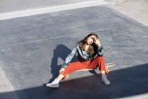 From above of young woman in stylish clothes touching forehead while sitting on longboard and looking at camera — Fotografia de Stock