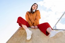 Low angle of female teen in trendy clothes and gumshoes looking at camera from concrete fence in back lit with blue clear sky — Fotografia de Stock