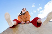 Low angle of female teen in trendy clothes and gumshoes looking at camera from concrete fence in back lit with blue clear sky — Stock Photo