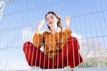 Знизу крутої жінки-підлітка в стильному одязі з бездротовими навушниками дивиться на камеру за металевим парканом в місті — стокове фото