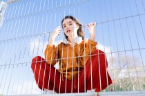 From below of cool female adolescent in stylish apparel with wireless headphones looking at camera behind metal fence in town — Photo de stock