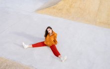 Сверху все тело молодая женщина в стильном наряде сидит в бетонном скейт-парке, глядя на камеру при солнечном свете — стоковое фото