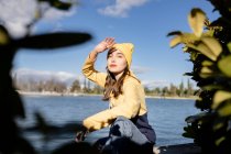 Жінка-підліток в стильному вбранні з макіяжем дивиться на камеру проти озера під блакитним хмарним небом — стокове фото