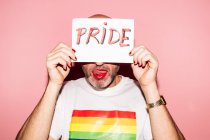 Rebellious bearded homosexual man with red lips and manicure making grimace with tongue out while showing and covering face with paper with Pride text against pink background — Fotografia de Stock