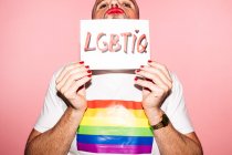 Rebellious bearded homosexual man with red lips and manicure making grimace while showing and covering face with paper with LGBTIQ text against pink background — Fotografia de Stock