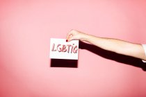 Crop unrecognizable homosexual person with manicure demonstrating white paper with LGBTIQ inscription against pink background — Photo de stock