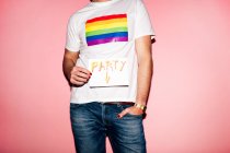 Crop unrecognizable homosexual guy in white t shirt with rainbow flag standing against pink background and showing paper with Party inscription — Stock Photo