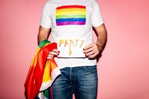 Crop unrecognizable homosexual guy in white t shirt with rainbow flag standing against pink background and showing paper with Party inscription — Fotografia de Stock