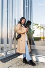 Повна довжина щасливої азіатської жінки в стильному одязі стоїть на вулиці і переглядає Інтернет на мобільному телефоні — стокове фото