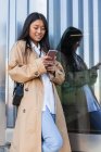 Glückliche Asiatin in stylischem Outfit steht auf der Straße und surft auf dem Handy im Internet — Stockfoto