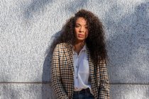 Pensive African American female in trendy outfit with curly hair looking at camera while standing on street near concrete wall — Fotografia de Stock