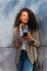 Афроамериканка в модном наряде с кудрявыми волосами, бродит по телефону, стоя на улице возле бетонной стены — стоковое фото