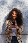 African American female in trendy outfit with curly hair browsing phone while standing on street near concrete wall — Stock Photo