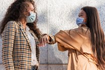 Side view of ethnic female friends in casual clothes with medical masks looking at each other while greeting with elbows — Stock Photo