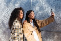 Ottimiste amiche etniche in abiti alla moda in piedi sulla strada della città e scattare selfie pur avendo pausa — Foto stock