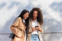 Оптимістичні етнічні друзі в модних вбраннях, що стоять на міській вулиці і спілкуються на мобільному телефоні, маючи перерву — стокове фото