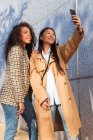 Optimistic ethnic female friends in trendy outfits standing on city street and taking selfie while having break — Photo de stock