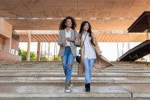 Delighted female friends with toothy smiles wearing trendy clothes looking away while walking together stairway in town — Stock Photo