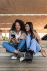Optimistische ethnische Freundinnen in trendigen Outfits sitzen auf der Straße und chatten auf dem Handy — Stockfoto