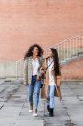 Захоплені друзі-жінки з зубними посмішками носять модний одяг, дивлячись далеко під час прогулянки разом у місті — стокове фото